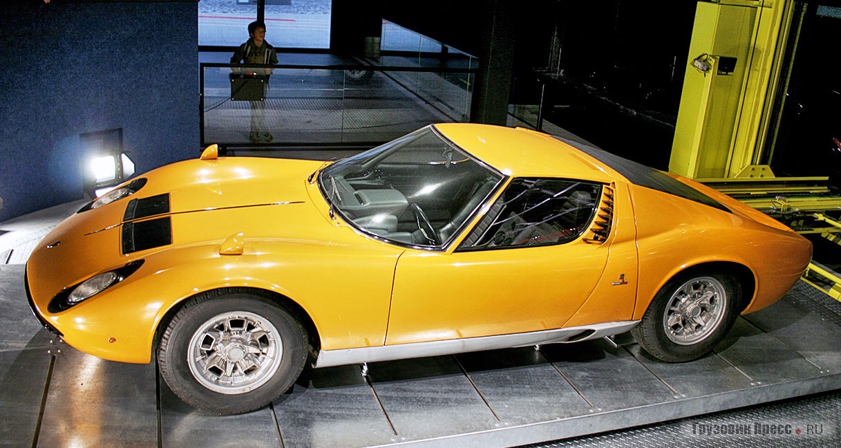 [b]Lamborghini Miura[/b] (1968 г.) фирменного апельсинового цвета. Выпускалась с 1968 по 1971 г. Сейчас это двухдверное итальянское купе на двоих – большая редкость. Автомобиль имел габариты 4360х1780х1060 мм, колёсную базу 2504 мм и дорожный просвет 130 мм. Продольно установленный V-образный 12-цилиндровый бензиновый двигатель объёмом 3929 cм<sup>3</sup> обладал мощностью 370 л.с., развивал максимальный крутящий момент 388 Н∙м. В смешанном цикле мотор потреблял 21 л на 100 км, отсюда и 90-литровый топливный бак. Время разгона с 0 до 100 км/ч занимало 6,7 с, потолок скорости достигал 276 км/ч
