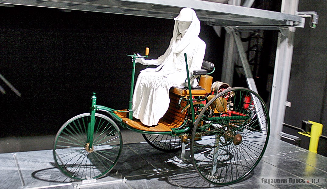 [b]Benz Patentwagen[/b] (1886 г.). Копия первого в мире автомобиля Карла Бенца образца 1886 г. с имитацией фигуры его супруги Берты, совершившей знаменитый автопробег. Одноцилиндровый двигатель развивал мощность всего 1 л.с. При массе 260 кг скорость не превышала 15 км/ч. Покупатели приняли новинку холодно. Тогда Берта Бенц без ведома мужа взяла автомобиль и с двумя старшими сыновьями совершила поездку из Мангейма в Пфорцгейм. В течение одного дня 5 августа 1888 г. она преодолела 106 км – огромное по тем временам расстояние. По пути они покупали бензин в аптеках и чистили тормозные колодки. Пришлось обращаться к кузнецу, чтобы наладить приводную цепь. Подвязка на женском чулке пригодилась для изоляции в системе зажигания, а заколка для волос – при чистке засорившегося бензопровода. Из-за слабого мотора на подъёмах приходилось толкать автомобиль в гору, что позже навело инженера на мысль о необходимости создания коробки передач